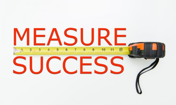 A yellow tape measure with the words "MEASURE SUCCESS" written on the side in black capital letters. The tape measure is extended to 11 inches.